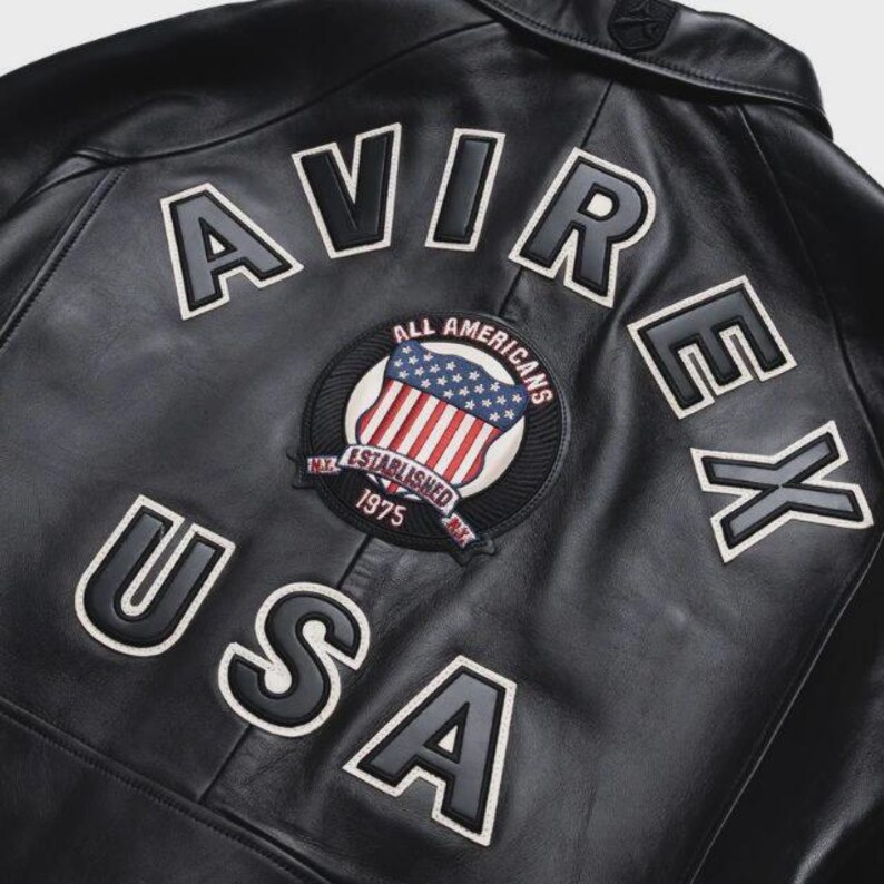 May include: Black leather jacket with the word "AVIREX" and "USA" in white lettering. A circular patch features the American flag and the words "ALL AMERICANS" and "ESTABLISHED 1975". The jacket has a classic design.