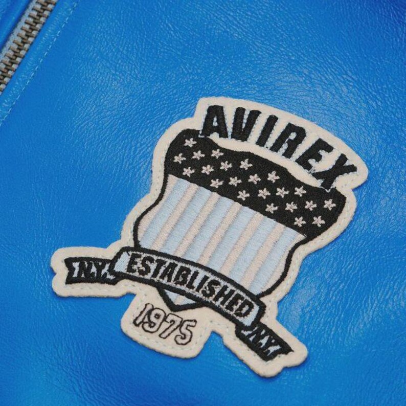 May include: Close-up of a bright blue leather jacket with a white and black patch. The patch features the word "AVIREX" above a shield design with stars and stripes, and the words "ESTABLISHED 1975 NY".