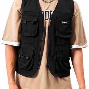 May include: A black utility vest featuring multiple front pockets with flaps and a central zipper closure. A small rectangular patch on the upper left chest pocket displays the white text So what! in a sans-serif font. The vest is sleeveless.