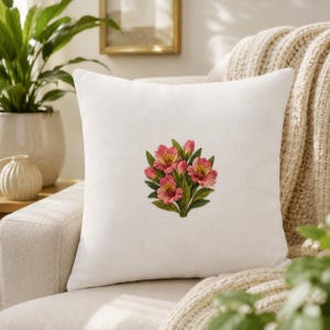May include: A square white scatter cushion featuring a central embroidered design of a cluster of pink alstroemeria flowers with green leaves. The fabric has a smooth, matte texture, and the floral arrangement displays detailed petals in shades of pink.