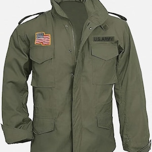 Könnte beinhalten: Eine olivgrüne Feldjacke mit hohem Kragen, langen Ärmeln und vier Vordertaschen mit geknöpften Klappen. Auf der rechten Brust befindet sich ein rechteckiger Aufnäher der amerikanischen Flagge, links ein schwarzer Aufnäher mit U.S. ARMY Text.