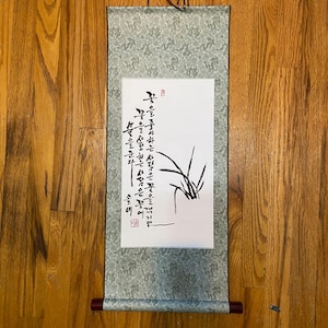 May include: A traditional Asian scroll with calligraphy and a botanical illustration. The scroll features a light green patterned fabric with wooden dowels at the top and bottom. The artwork includes black ink calligraphy and a drawing of a plant.