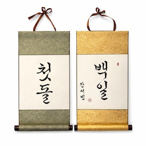 May include: Two Korean traditional scrolls. One is green and the other is gold. Each scroll has black calligraphy on a white background. The scrolls have brown wooden dowels at the bottom and brown ribbon at the top.