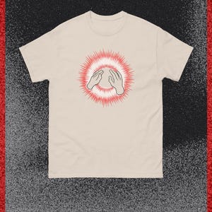 以下が含まれることがあります： クリーム色のTシャツ。2つの手を合わせたグラフィックが特徴で、赤いバーストデザインで囲まれています。柔らかい素材でできており、半袖です。