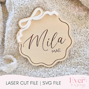 May include: A wooden name tag with a scalloped edge and a white bow accent. The name "Mila" is written in a flowing script, with "MAE" below it. The text "LASER CUT FILE | SVG FILE" is at the bottom.