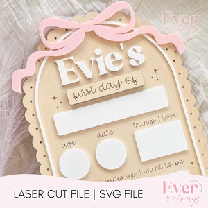 以下が含まれることがあります： ピンクのリボンが付いた木製のサインで、白文字で「Evie's」と表示されています。「first day of」、年齢、日付、「things I love」のセクションがあります。このサインは、子供のマイルストーンを記念するために設計されています。