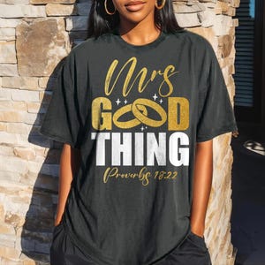 May include: Black t-shirt with gold and white text that reads "Mrs. Good Thing" with wedding rings and "Proverbs 18:22". The shirt is a relaxed fit.