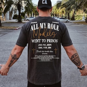 May include: Dark gray t-shirt with the text "ALL MY ROLE Models WENT TO PRISON" in white and gold lettering. The shirt also features the names Jesus, Paul, Joseph, Daniel, Peter, and John. A black baseball cap with "CA" is worn.