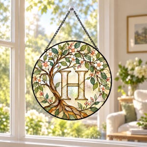 May include: A circular stained glass suncatcher featuring a tree with green leaves and pink blossoms surrounding a large, central letter H. The piece is framed in black metal and hangs from a black chain attached to a clear suction cup.
