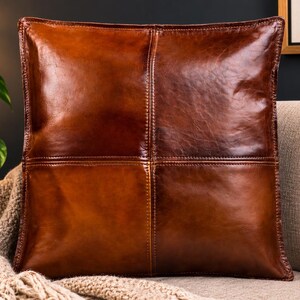 Könnte beinhalten: Ein quadratisches Zierkissen aus sattem, dunkelbraunem Leder. Die Oberfläche weist ein markantes Vier-Felder-Design auf, das durch vertikale und horizontale Nähte entsteht, welche das Kissen in gleichmäßige Quadranten mit glatter Oberfläche unterteilen.