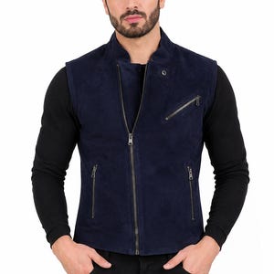 Può includere: Un gilet in camoscio blu navy scuro con colletto alla coreana, cerniera centrale a tutta lunghezza, due tasche inferiori con zip e una tasca sul petto inclinata con zip. Il capo è indossato sopra una maglia nera a maniche lunghe e pantaloni.
