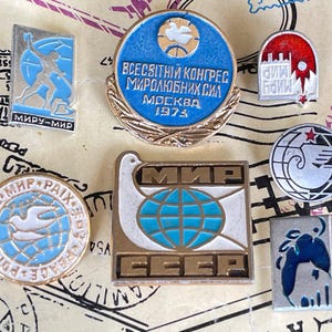 Puede incluir: Una colección de siete pines de solapa de metal vintage con varios diseños, incluyendo un pin circular azul con el texto "ВСЕСВІТНІЙ КОНГРЕС МИРОЛЮБНИХ СИЛ МОСКВА 1973" y un pin cuadrado con una paloma blanca, un globo terráqueo azul y el texto cirílico "МИР" y "СССР".
