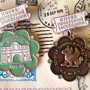 May include: Two metal souvenir pins with white scroll-shaped headers featuring Cyrillic text. The left pin is a green flower shape with a white building illustration and the text 1706-1711. The right pin is a dark brown flower shape with a gold griffin.