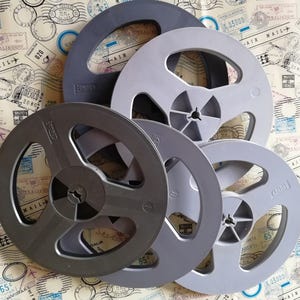 Può includere: Una collezione di quattro bobine circolari in plastica per pellicole in varie tonalità di grigio e nero. Le bobine presentano un mozzo centrale con raggi e aperture, alcune con scritte in rilievo sulla superficie in plastica.