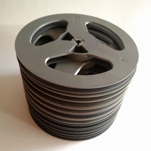 Può includere: Una pila di bobine circolari in plastica grigia. Ogni bobina presenta un design a tre razze con un mozzo centrale e una piccola apertura circolare. Le bobine sono impilate verticalmente, creando una forma cilindrica con bordi visibili.