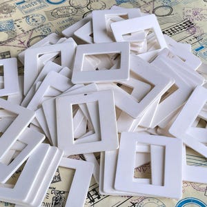May include: A large collection of square white plastic slide mounts, each featuring a central rectangular opening. These empty frames are stacked in a disorganized pile, showcasing their uniform matte finish and rigid construction.