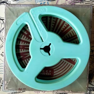 Può includere: Una bobina di pellicola in plastica verde menta chiaro con un design a tre razze e un foro centrale triangolare. La bobina presenta marcature in rilievo per 9,5 cm e 19 cm. All'interno, una striscia di pellicola mostra motivi rossi e bianchi con il testo CCCP MOSKVA.