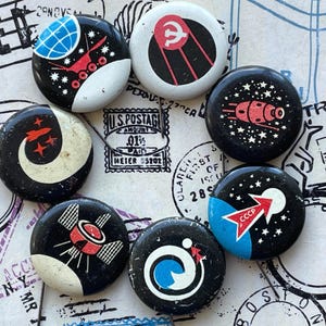 Puede incluir: Una colección de siete chapas redondas con motivos espaciales. Los diseños incluyen un satélite rojo, un cohete, un globo terráqueo y el texto CCCP. Cada chapa tiene un fondo negro o blanco con ilustraciones espaciales de colores.