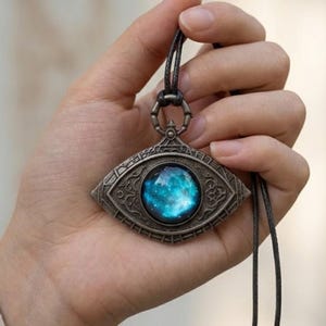 Può includere: Un ciondolo a forma di occhio con una cornice in metallo color bronzo antico e dettagli in filigrana. Il centro contiene un cabochon circolare che mostra un vivace motivo a nebulosa blu e nero. Il ciondolo è appeso a un cordino scuro.