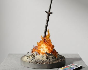 Könnte beinhalten: Eine dekorative Lampe mit einem dunkelgrauen Schwert, das in einem Haufen durchscheinender orangefarbener Flammen auf einem runden Sockel steckt. Der Sockel enthält kleine graue knochenartige Fragmente und eine rechteckige Fernbedienung.