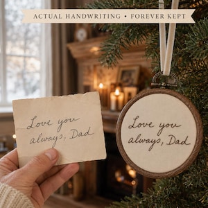 Pode incluir: Um ornamento redondo de madeira para bordado com tecido off-white apresentando o texto bordado em preto "Love you always, Dad" pendurado por uma fita branca, ao lado de um papel com o mesmo texto manuscrito em tinta preta.