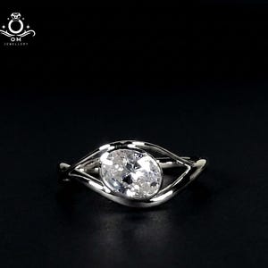May include: A silver ring with a large, oval-cut, clear gemstone. The ring's band has a unique, flowing design that curves around the gemstone. The ring is set against a black background, highlighting its brilliance.