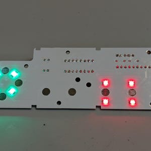 May include: A white printed circuit board featuring four illuminated green LEDs arranged in a circular pattern on the left side and four illuminated red LEDs arranged in a rectangular pattern on the right side, with various circular contact points.