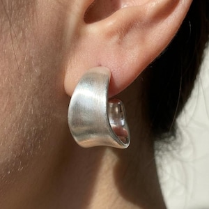 May include: A thick, curved silver hoop earring with a brushed matte finish. The earring has a wide, sculptural shape that tapers slightly at the ends, creating a smooth, continuous arc that sits securely against the earlobe with a polished interior.
