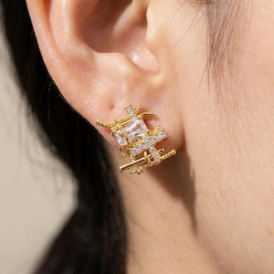 May include: A gold-toned stud earring featuring a complex, layered structure of thin metal bars and wire-like accents. The design incorporates a central rectangular clear gemstone and smaller pavé-set clear stones along the metallic framework.