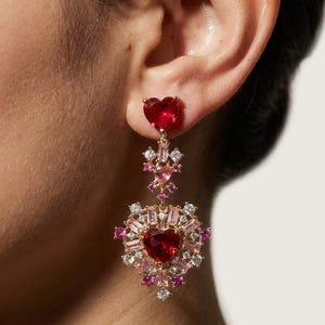 May include: A gold-tone drop earring featuring a heart-shaped red stone at the top, connected by a small floral-inspired link with pink and clear stones to a larger, ornate heart-shaped pendant set with red, pink, and clear baguette and round stones.