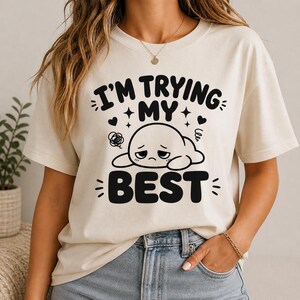 Pode incluir: Camiseta creme com o texto preto "I'M TRYING MY BEST" e um personagem de desenho animado triste. O design inclui corações e estrelas. É uma peça casual para o dia a dia.