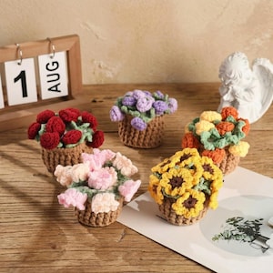 May include: A collection of handmade crochet flower arrangements in small woven baskets. The flowers are in various colors, including red, pink, yellow, and purple. A wooden calendar shows "1 AUG". A small white angel statue is in the background.