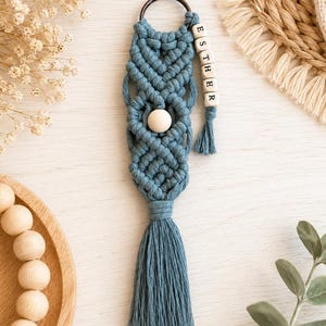 May include: A blue macramé keyring featuring intricate knotted patterns, a central round wooden bead, and a dangling strand of square wooden beads spelling the name ESTHER. The design is finished with a long, thick blue tassel and a metal ring.