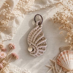May include: A macrame keyring in a light beige colour, shaped like a seashell with a spiral pattern. The design features a single smooth, round, off-white wooden bead attached to the lower section and a dark metal ring at the top for hanging.