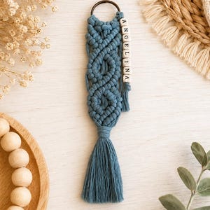 May include: A blue macrame keychain featuring a series of intricate knotted patterns, a dark metal ring, and a dangling strand of white square beads spelling the name ANGELINA, finished with a long, thick blue tassel at the bottom.