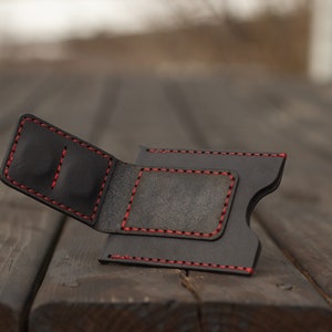 Puede incluir: Una cartera de cuero negro con costuras rojas. La cartera está abierta y tiene varias ranuras para tarjetas.