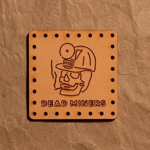 May include: A brown leather patch with a skull wearing a miner's hat and the text "DEAD MINERS".
