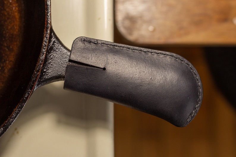 May include: A black cast iron skillet with a dark brown leather handle cover.