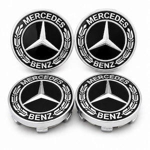May include: A set of four circular wheel centre caps featuring the Mercedes-Benz logo. Each cap has a black background with a silver three-pointed star in the centre, surrounded by a silver laurel wreath and the text Mercedes-Benz in silver.