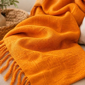 Puede incluir: Una manta tejida naranja con flecos. La manta está sobre un sofá de color neutro. En el fondo se ve una cesta tejida y un pequeño jarrón con vegetación.