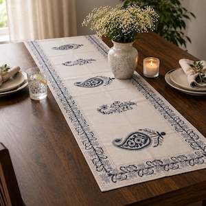 May include: A cream-colored table runner with a blue paisley pattern and border is displayed on a dark wooden table. The table is set with plates, glasses, a vase of flowers, and a lit candle, creating a dining setting.