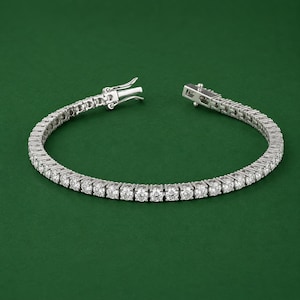 May include: A silver tennis bracelet with a row of round, clear gemstones. The bracelet is open, showing the clasp mechanism. The background is a solid, dark green color.