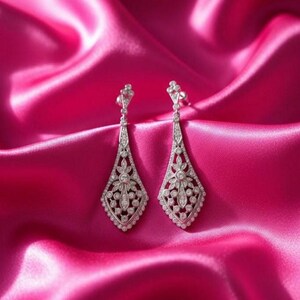 May include: A pair of silver-toned drop earrings with a floral and geometric design, adorned with numerous small, sparkling gemstones. The earrings are displayed against a vibrant pink satin background, creating a luxurious and elegant aesthetic.