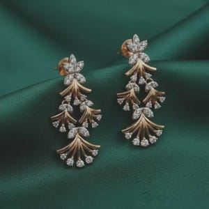 May include: A pair of gold-toned earrings with a floral design. Each earring features a flower-shaped stud and a cascading arrangement of leaf-like elements adorned with small, sparkling gemstones. The earrings are set against a dark green background.