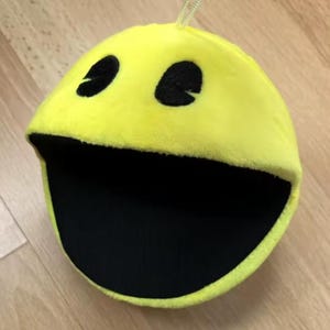 Può includere: Un peluche giallo di Pac-Man con occhi neri e una grande bocca aperta. Il peluche ha un anello nella parte superiore per appenderlo. Il peluche di Pac-Man è un personaggio classico dei videogiochi.