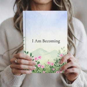 May include: A notebook with the words "I Am Becoming" on the cover. The cover has a watercolour design with a blue sky, green hills, and pink flowers. The notebook is held by a person.