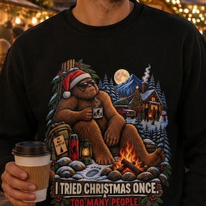 May include: A black crewneck sweatshirt featuring a graphic of a brown Bigfoot wearing a Santa hat and sunglasses, sitting by a campfire in a snowy forest. The text reads, I TRIED CHRISTMAS ONCE. TOO MANY PEOPLE. The design includes a cabin and moon.