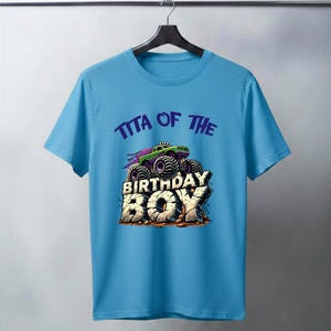 May include: A light blue t-shirt featuring a graphic of a green and purple monster truck with the text "TITA OF THE BIRTHDAY BOY". The design has a distressed, stone-like appearance for the text "BIRTHDAY BOY".