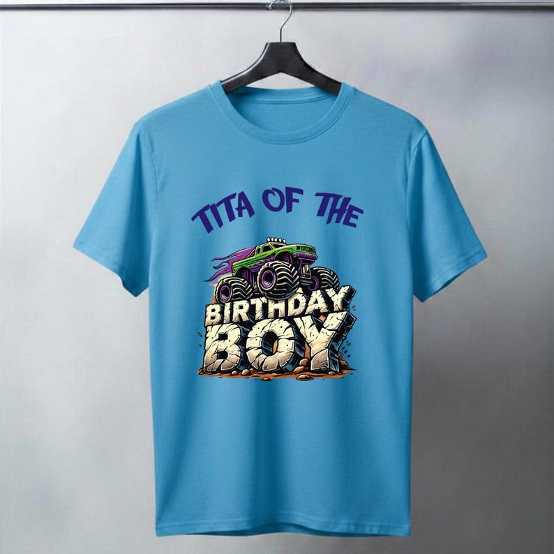 May include: A light blue t-shirt featuring a graphic of a green and purple monster truck with the text "TITA OF THE BIRTHDAY BOY". The design has a distressed, stone-like appearance for the text "BIRTHDAY BOY".