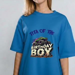 May include: Blue t-shirt with a graphic of a monster truck and the text "Tita of the Birthday Boy". The monster truck is green and purple. The text "Birthday Boy" is in a bold, blocky font.